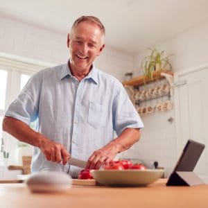 Retired Man Making Meal In Kitchen Asking Smart Speaker
